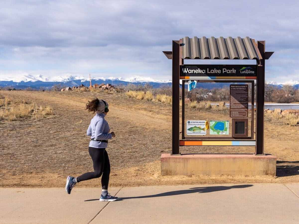 Woman jogging at Waneka Lake Park with park sign and lake and mountains in the background for article Lafayette Colorado for newcomers moving to Denver area.