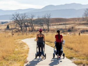 2 women in active wear pushing strollers down walking trail at Chatfield State Park with mountains in the background for article Best Places to Raise a Family in Colorado for newcomers moving to Denver area.