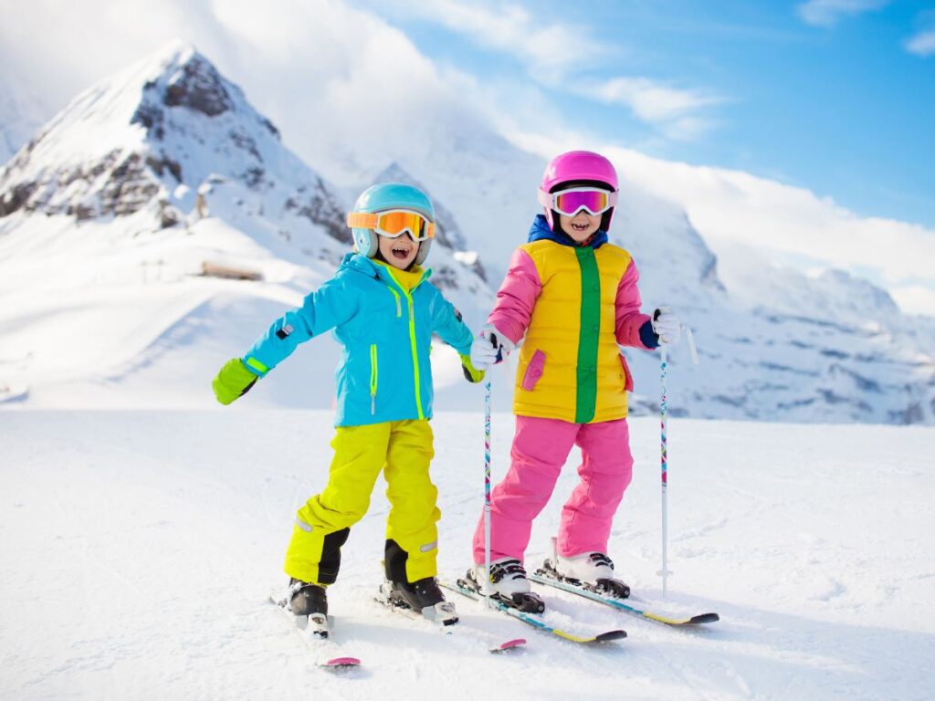 2 kids dressed in yellow and pink snow pants and blue and pink snow jackets and helmets with goggles posing for camera on snow skis in Colorado mountains for article Outdoor Adventures With Colorado Kids