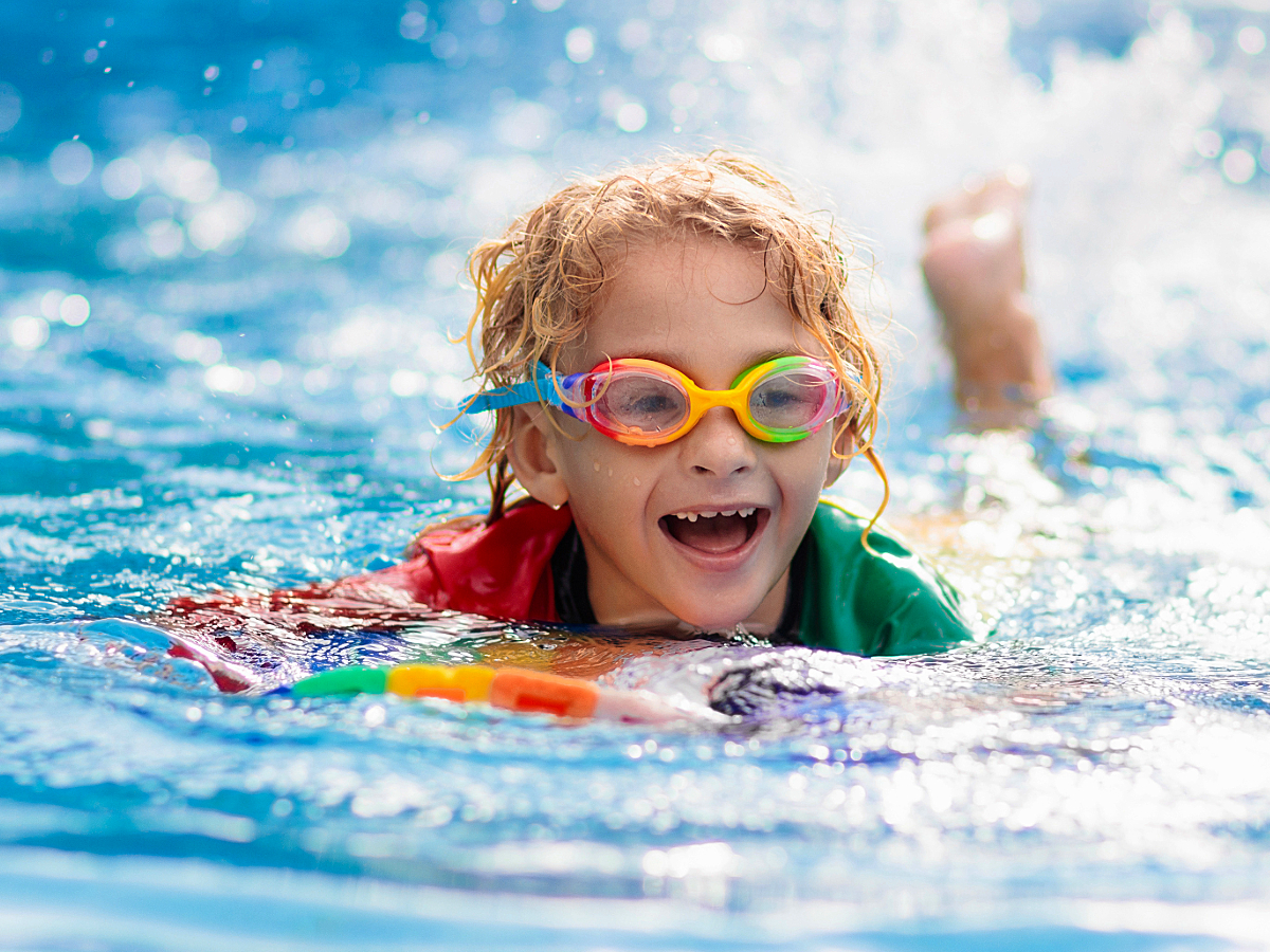 Kid with rainbow goggles on and a rainbow kick board splashing, learning to swim and smiling for article Top 10 Swimming Schools for Kids in Denver: A Guide for Newcomers moving to Denver.