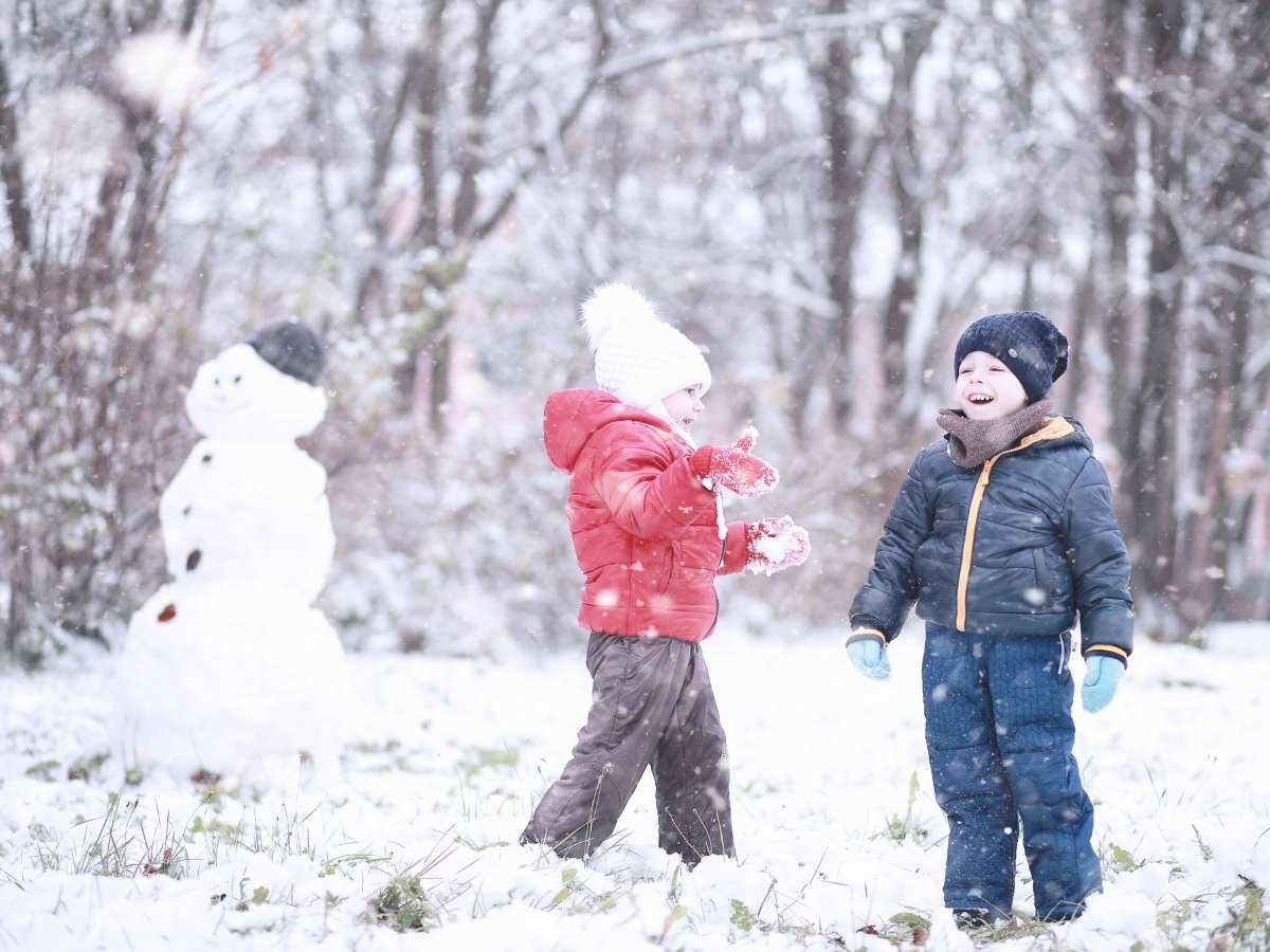2 kids bundled up in snow gear playing outside in the snow with a snowman in the background for article Keeping Colorado Kids Safe and Warm During the Winter for newcomers moving to Colorado.