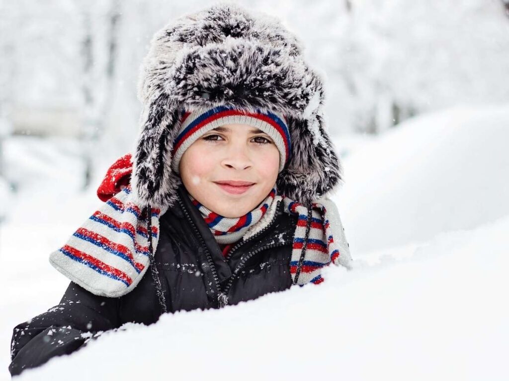 Boy wrapped up in coat, scarf , fuzzy hat surrounded by snow for article Children's Hospital Colorado: Health During the Holidays in Denver for newcomers moving to Denver.
