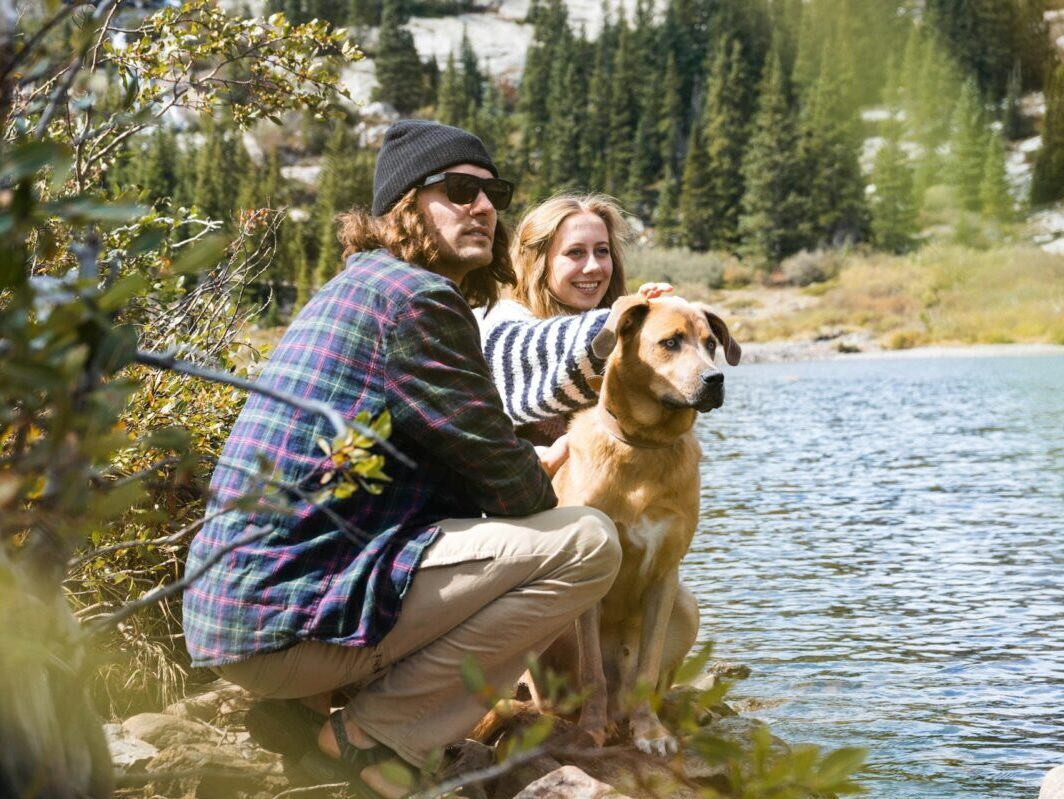 man in a purple and blue shirt, and woman in a blue and white striped shirt, petting down and kneeling in front of water.