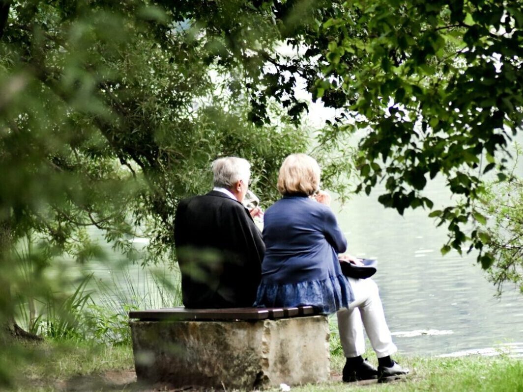 A senior couple dressed in nice clothes, drinking a glass of wine and eating lunch while sitting on stone looking out the water in daylight