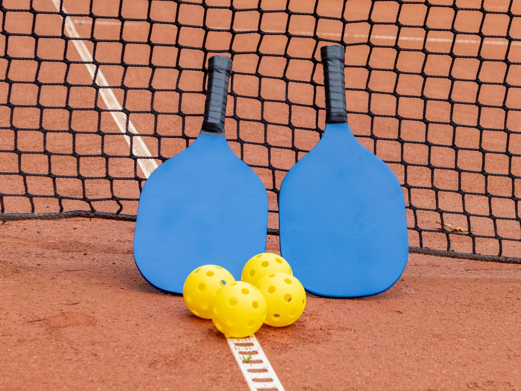 two pickleball rackets and four yellow pickleballs are standing upright along a net on a court