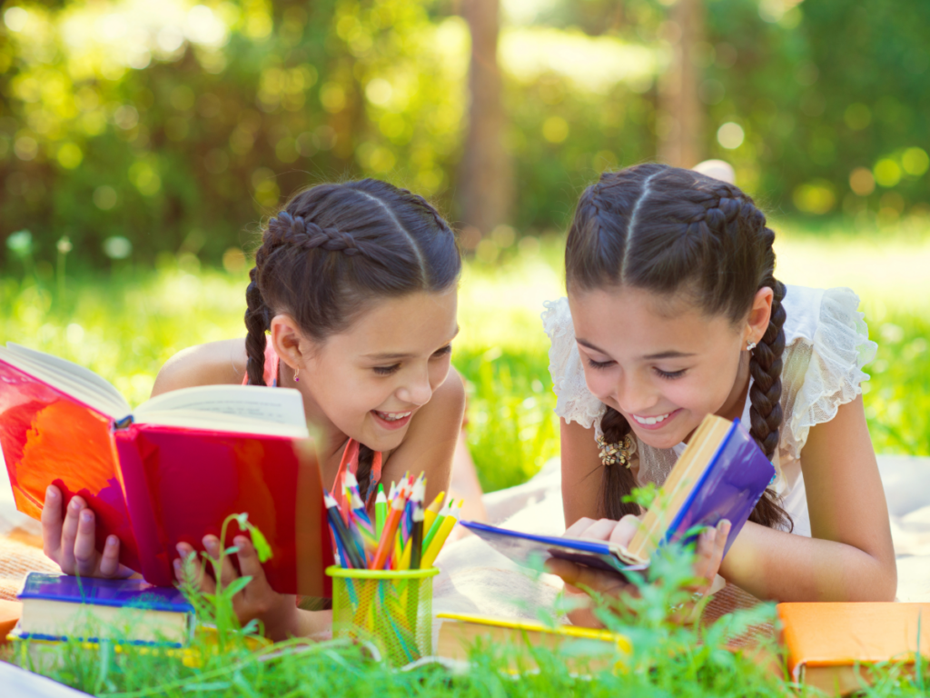 2 Young girls laying on the ground outside reading books.