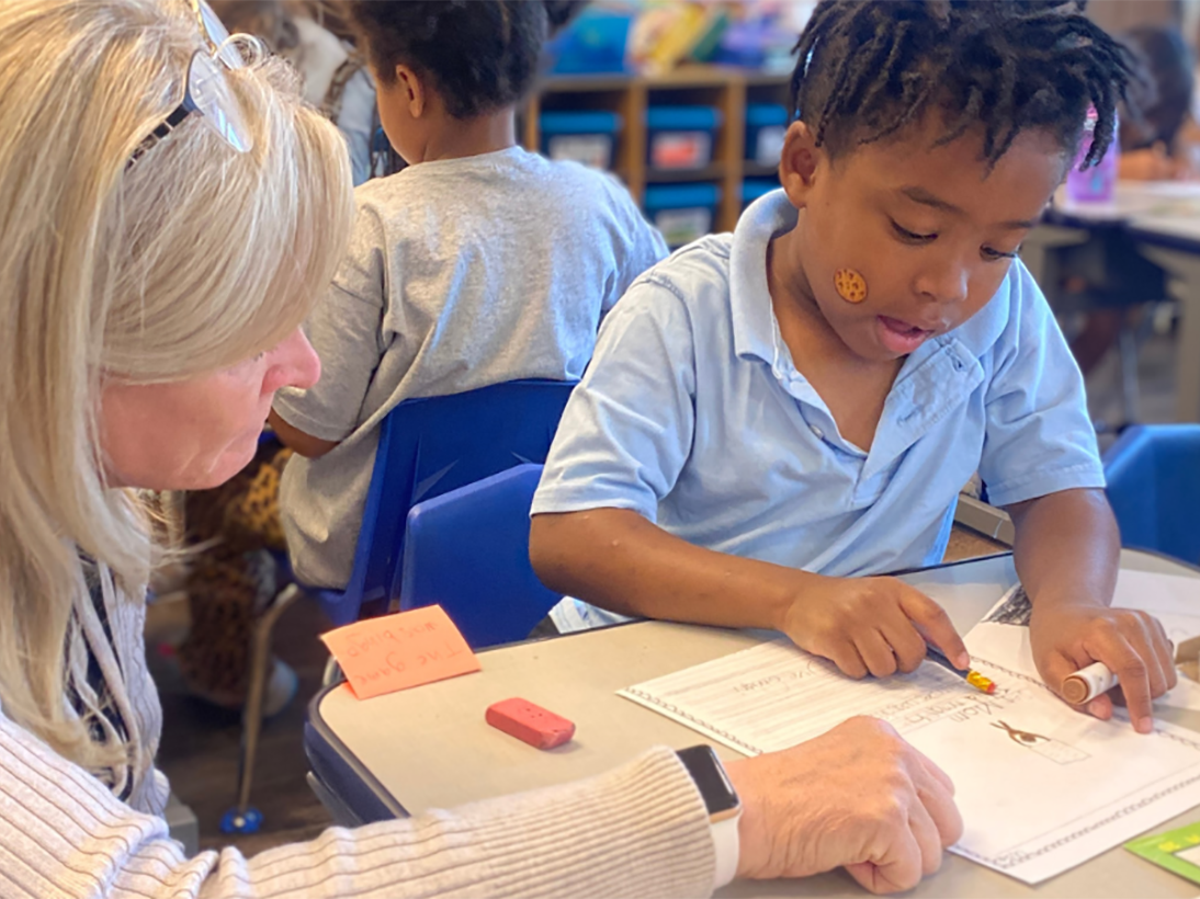 student in blue shirt sitting at desk with paper and pencil while teacher looks on