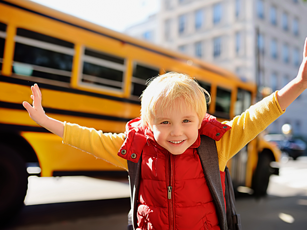 Young boy in red puffy best and backpack with arms outstretched smiling in front of school bus.