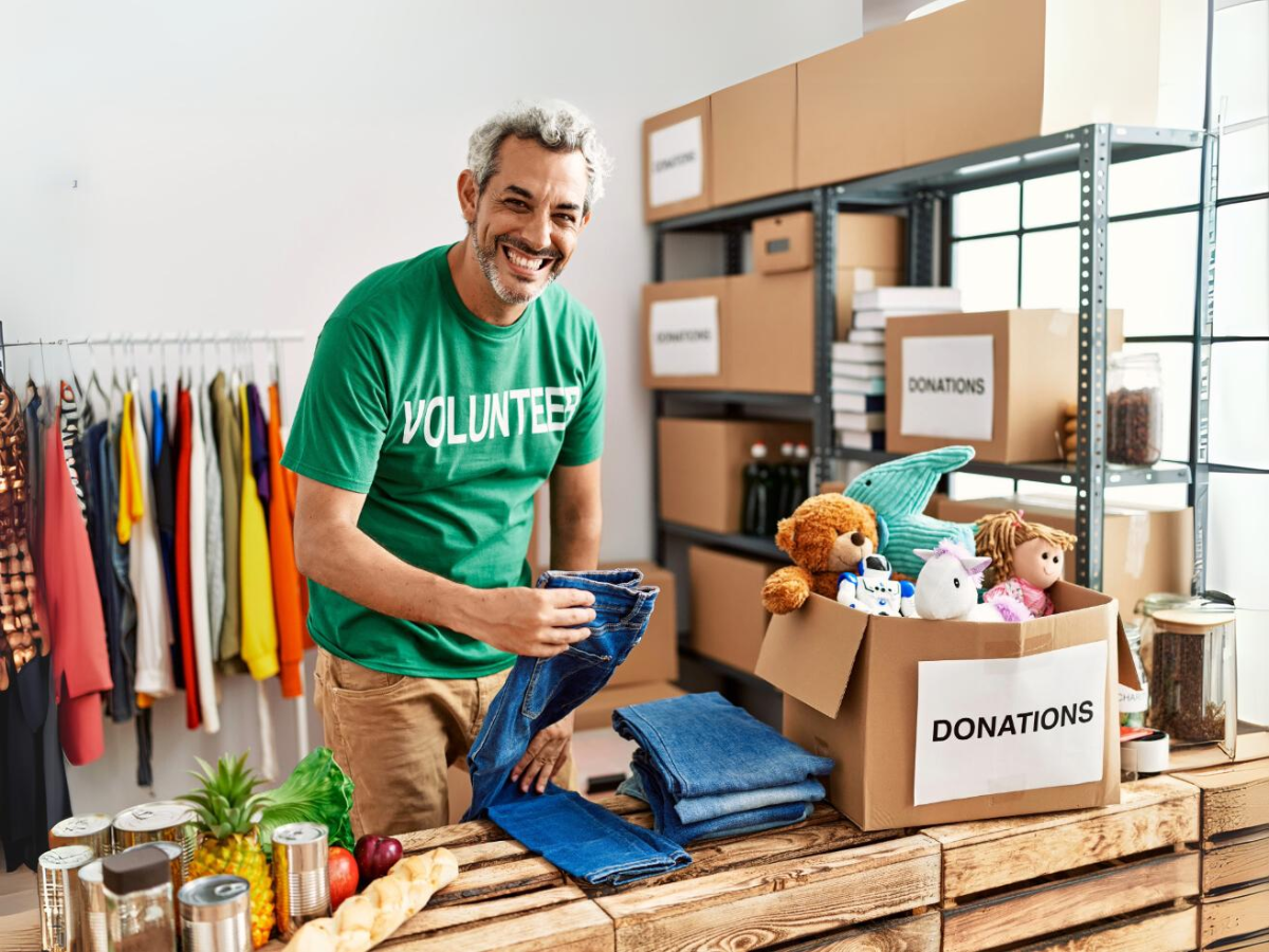 Senior man in green volunteer shirt folding denim with donation boxes around him.