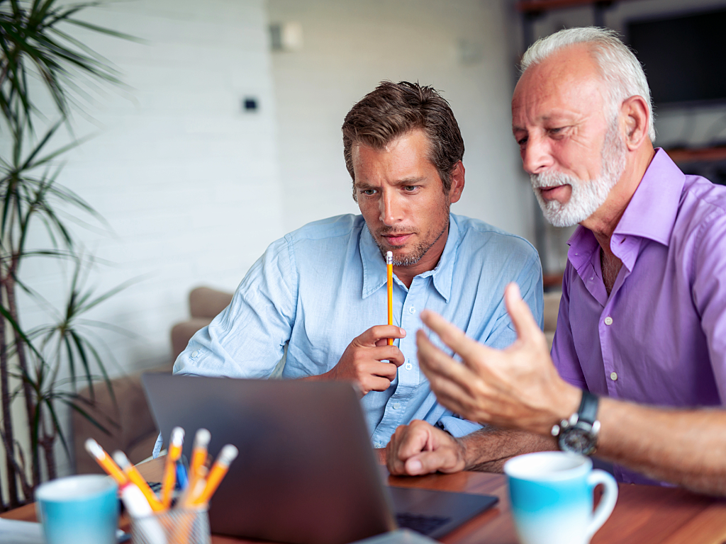 Older gentleman mentoring younger professional at a table with laptop in front of them.