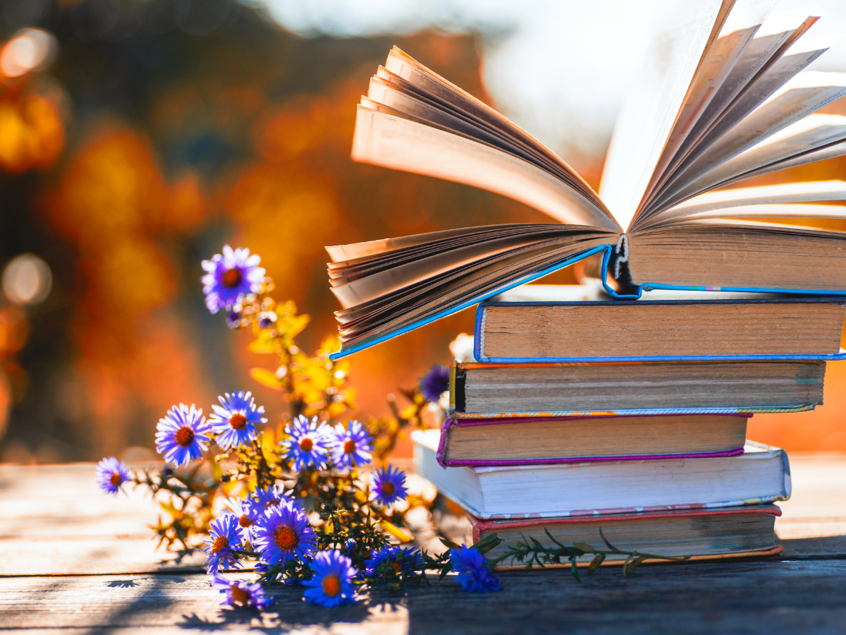 Stack of books on a table outdoors with flowers next to them.