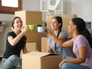 3 female roommates sitting on floor drinking coffee with moving boxes around them for article on Finding Roommates in Denver.