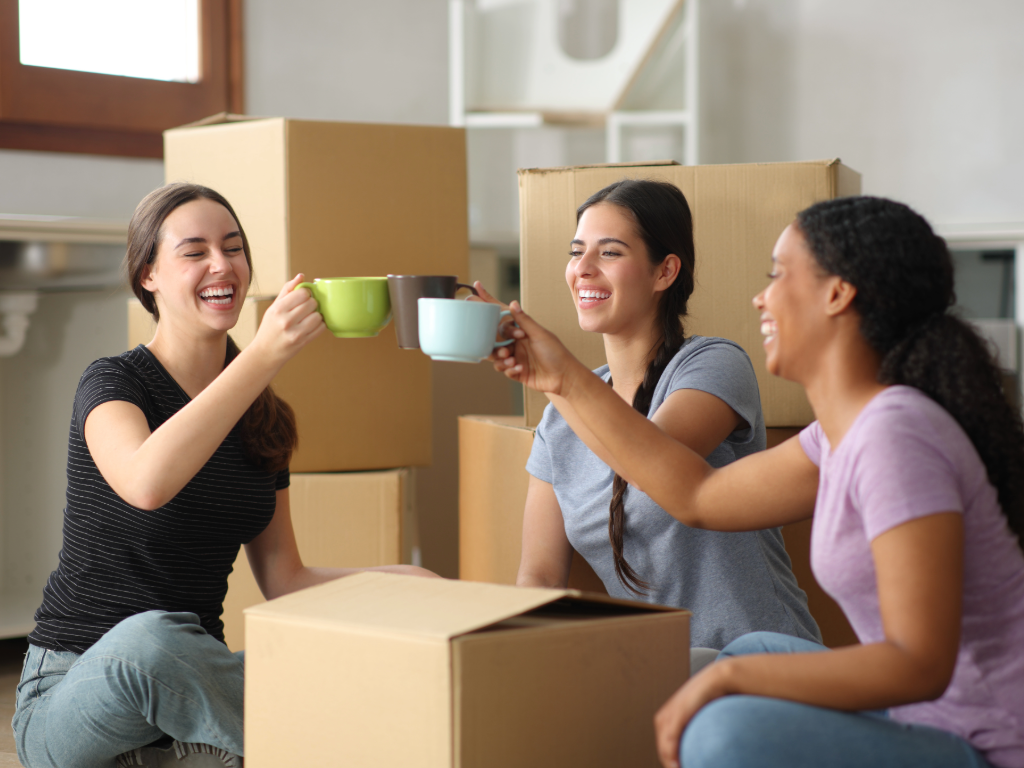 3 female roommates sitting on floor drinking coffee with moving boxes around them.