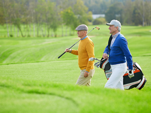 2 men walking side by side on hilly golf course. 1 is carrying golf club over shoulder, the other is carrying golf bag.