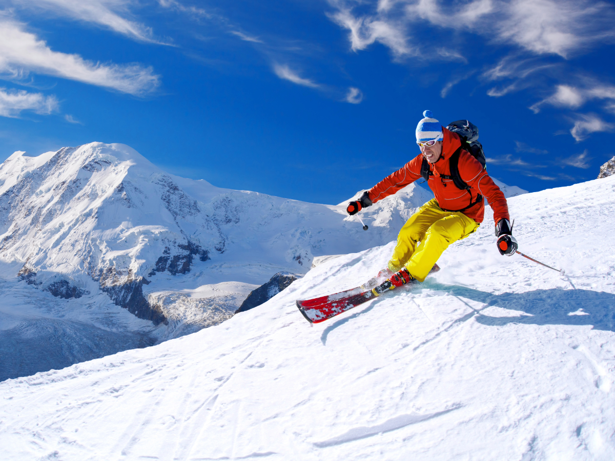 Colorado skier in red jacket and yellow pants snow skiing with blue sky and snow covered mountains in background for article Colorado Ski Season Opening Dates for 2024-2025.