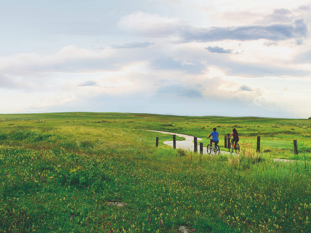 2 people riding bikes away from camera on bike path through green rolling hills for article Aurora, Colorado: The World in a City for newcomers moving to Denver.