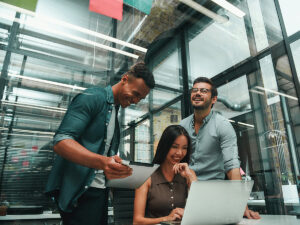 1 Female sitting at computer with 2 male coworkers standing behind her and collaborating with her in modern technology office environment for article Top 15 Denver High Technology and IT Companies for newcomers moving to Denver.