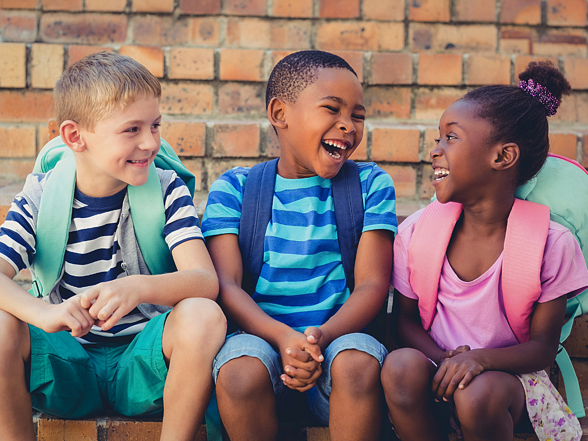 3 kids sitting in front of brick wall with backpacks on their backs, smiling and laughing for article Denvers Best School Districts for Newcomers: Top 8 Ranked for newcomers moving to Denver.