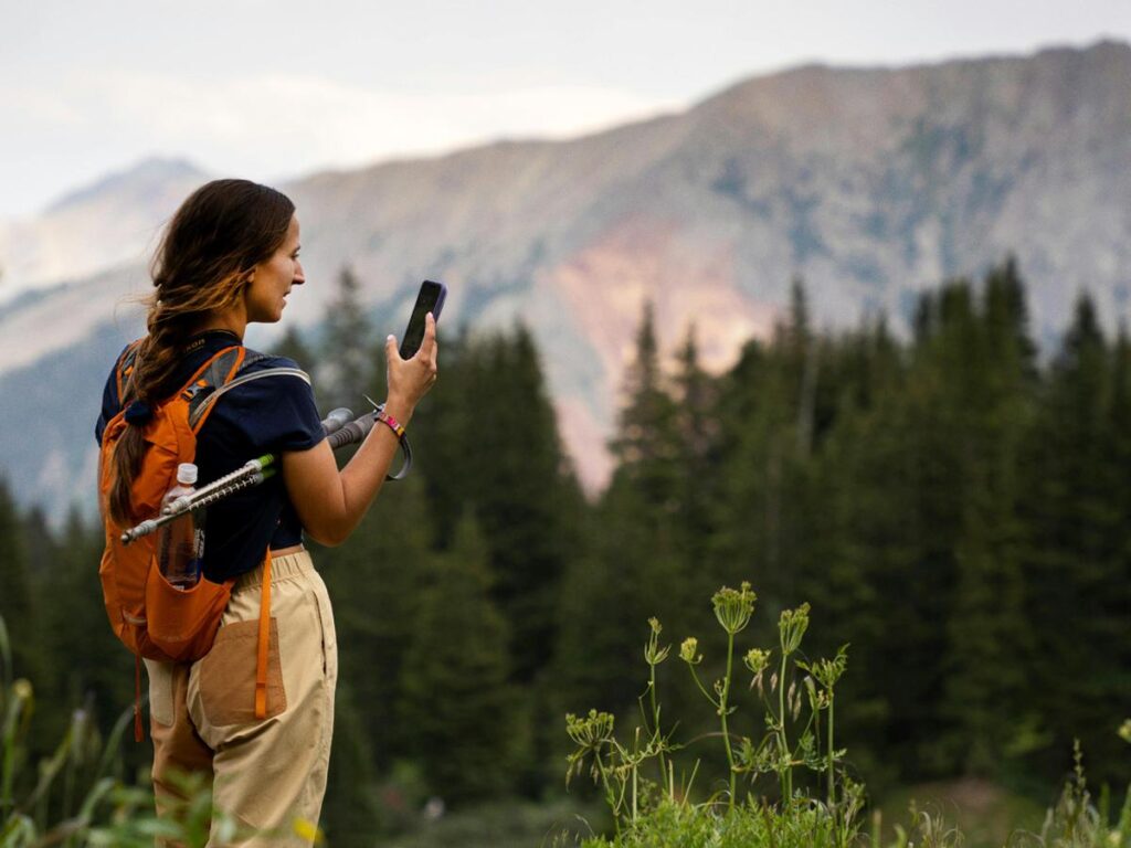 Woman holding phone out in hand while hiking in Colorado with orange backpack for article Credit Union of Colorado. Honestly, Good. for newcomers moving to Colorado.