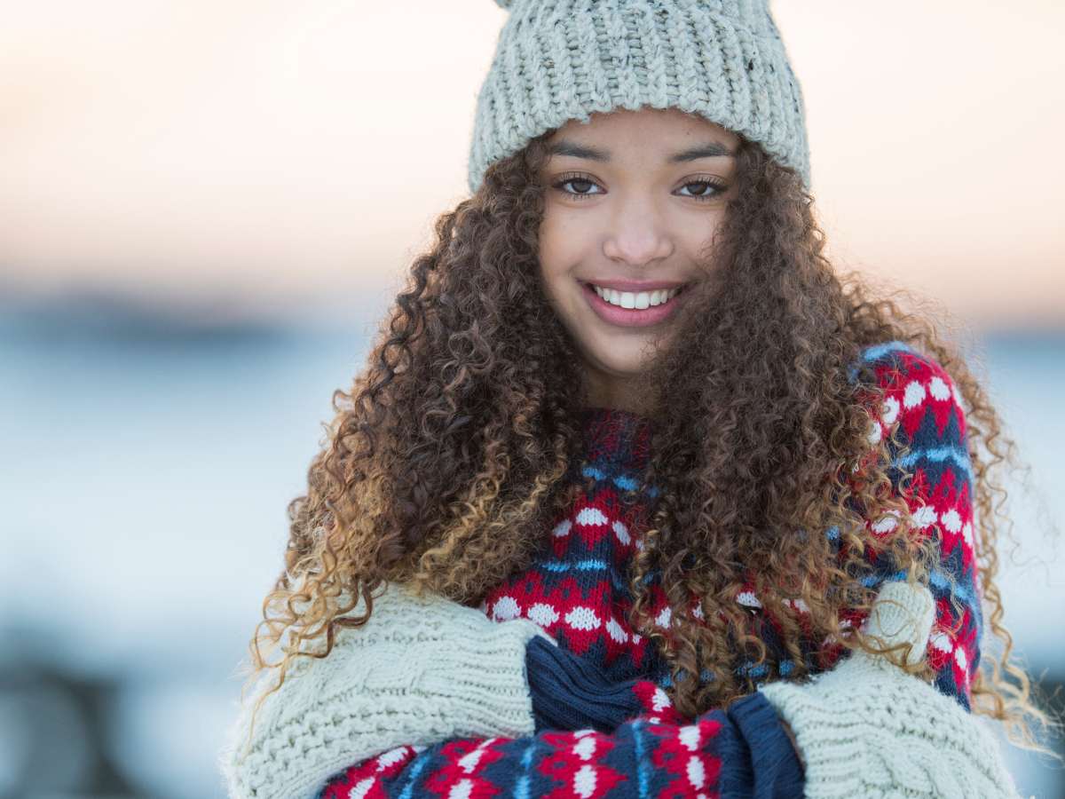 Close up of young woman with long curly hair with sweater, hat and gloves with armed crossed smiling at camera for article Women's Health in Denver: 14 Popular Resources for newcomers moving to Denver.