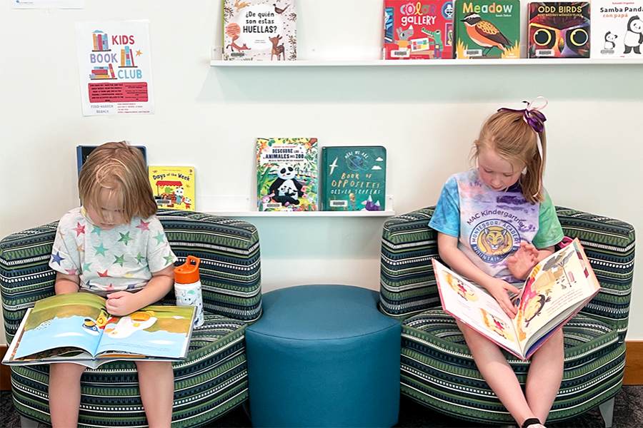 2 girls sitting on fun striped modern chairs with book shelves on walls behind them. The young girls are each reading a book for article Montessori Academy of Colorado for newcomers moving to Denver.