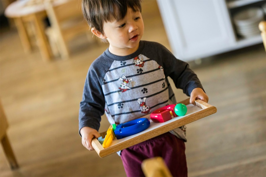 A boy walking with a tray of educational toys on a framed tray for article Montessori Academy of Colorado for newcomers moving to Denver.