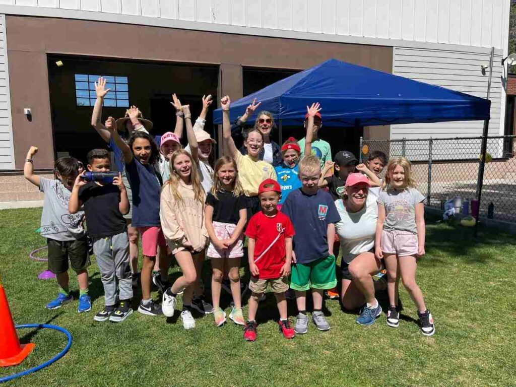 Group of students and teachers posing and cheering outdoors by a blue pop up tent for article The Joy Factor: How Montessori School of Denver Helps Kids Thrive for newcomers moving to Denver.