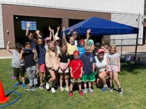 Group of students and teachers posing and cheering outdoors by a blue pop up tent for article The Joy Factor: How Montessori School of Denver Helps Kids Thrive for newcomers moving to Denver.