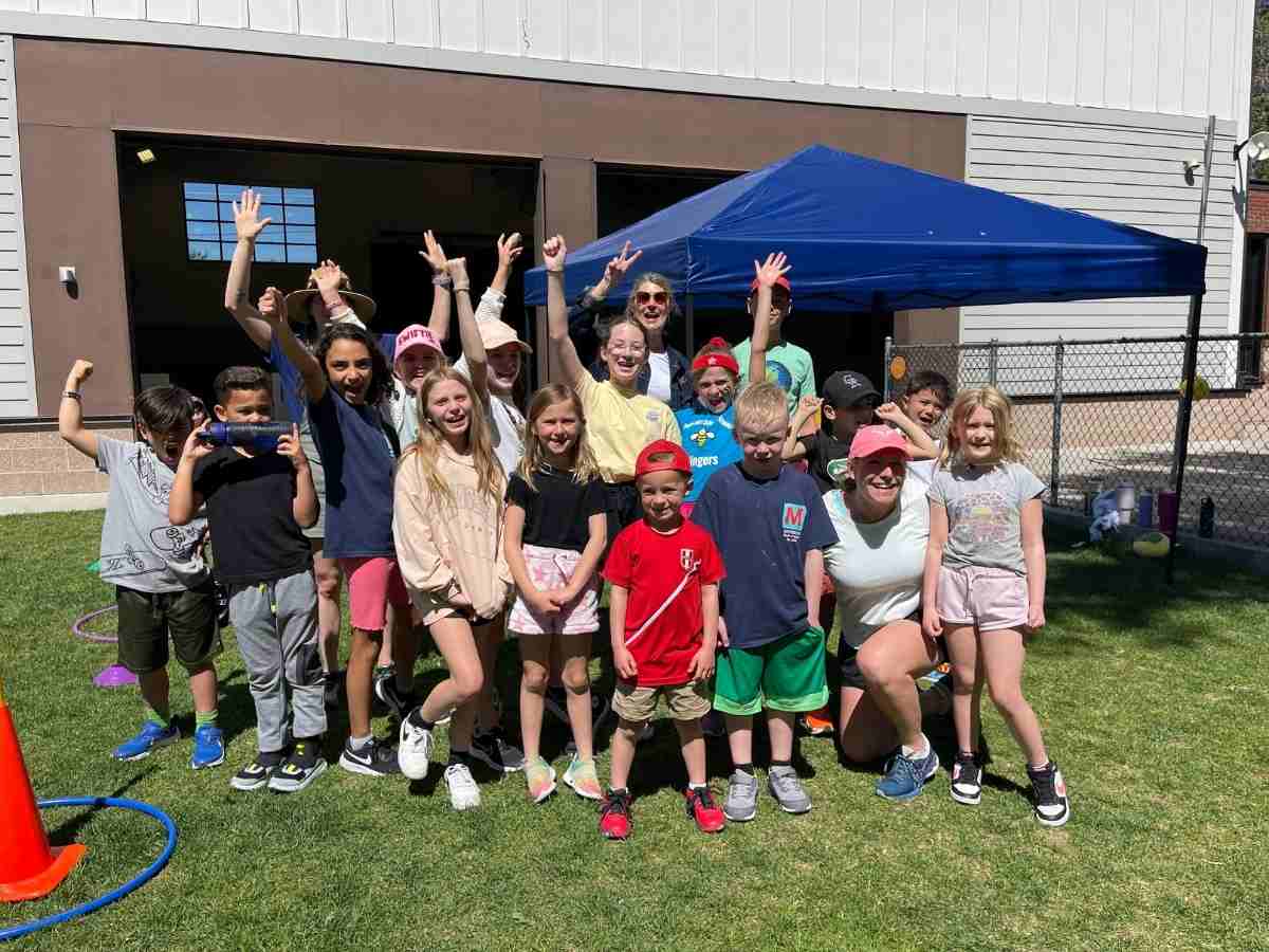 Group of students and teachers posing and cheering outdoors by a blue pop up tent for article The Joy Factor: How Montessori School of Denver Helps Kids Thrive for newcomers moving to Denver.