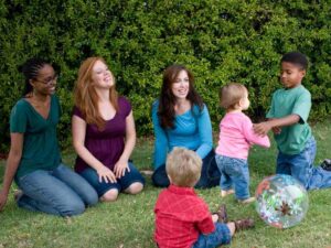 3 Moms with kids sitting outside watching their children play in the grass with a ball for article Denver Parenting Groups and Playdates for newcomers moving to Denver.