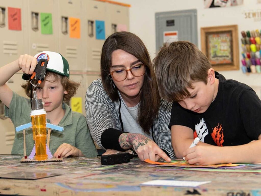 Teacher sitting at table in between 2 students helping with school projects for article Denver Academy for newcomers moving to Denver.