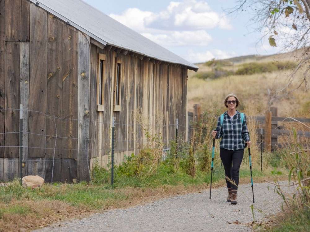 Woman hiking with walking sticks on trail by old barn and hills in background for article RidgeGate.